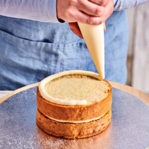 afbeelding van persoon met spuitzak met botercrème die biscuuit taart vult